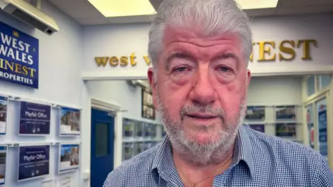 Neil Evans in is estate agency offices with home sales listing and the business logos on the walls behind him