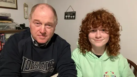 Heledd and her father Ifan looking at the camera. Ifan is on the left and is wearing a blue jumper that has Lonsdale written in block letters. He has a receding hairline. Heledd is on the right and has a pale green jumper on and long ginger curls.