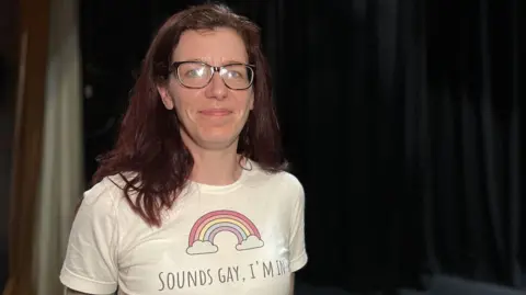 Kaye Nicholson is a woman with shoulder-length red hair and glasses. She is smiling at the camera and is wearing a white T-shirt with a rainbow on the front, bearing the slogan: "Sounds gay, I'm in."