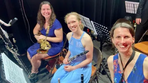 Liz Oughton Liz is sitting next to two women and they are all wearing blue outfits and holding instruments