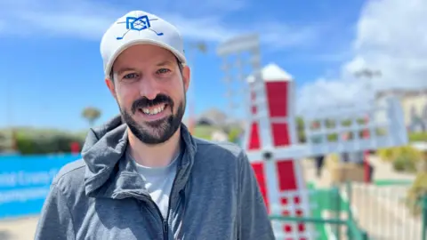 A man wearing a white hat with a blue logo and grey light jacket is standing at a mini golf course. 