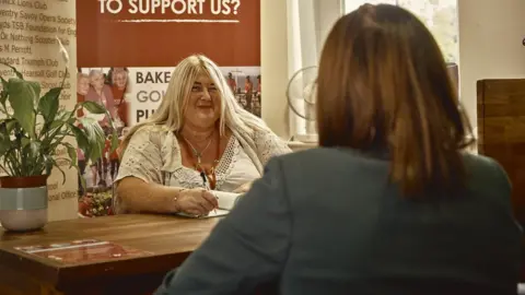 Myton Hospice Claire McDowell at her desk speaking to another person. She has blonde hair and with a big smile on her face is holding a pen and taking notes. 