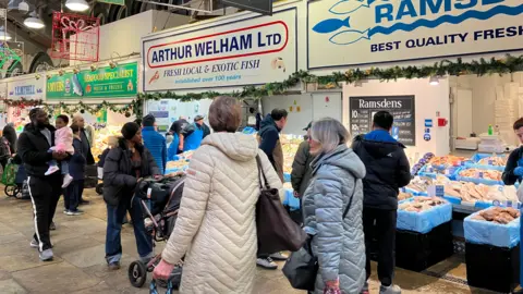 Shoppers wearing coats milling about some market stalls. Fishmongers can be seen in the background.