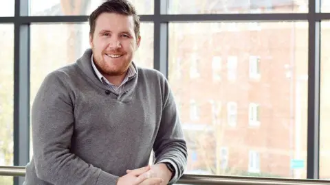 Philip Clarke A man in a grey sweater with a button-up collar leans casually against a handrail in front of a large glass window. Through the hazy outdoor light a block of flats is visible.