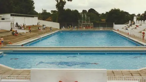Broomhill Pool Trust Broomhill lido pictured in 1969. Blue tiles in the shape of a whale can be seen at the bottom of the pool. There are people sitting around the edge of the pool.
