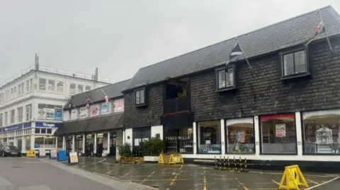 The two buildings which make up Trago Mills in Falmouth. One is wooden two-storey building and the other is a three-storey building along Arwenack Street. There is a hatched area in front of the wooden building and it has the nations' flags flying. 