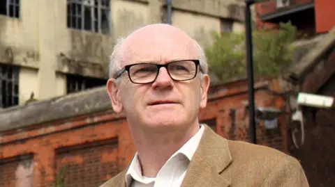 Ipswich Borough Council Neil MacDonald looks away from the camera. He is largely bald and is wearing a brown jacket with a white shirt underneath.