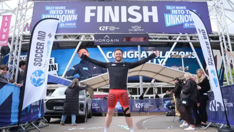 PA Singer Olly Murs crosses the finish line with his hands in the air wearing orange shorts and a black top
