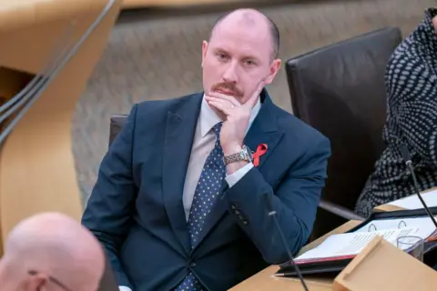 PA Media Neil Gray wears a dark suit and sit at a desk in the Scottish parliament chamber watching the first minister John Swinney speak.