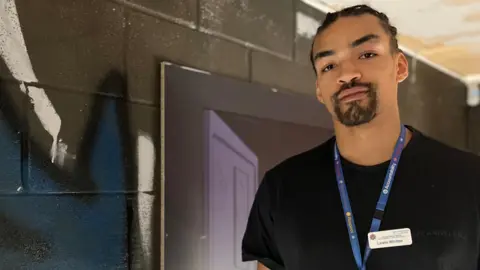 Lewis Whitter is stood in front of a graffiti covered wall. He is looking at the camera and wearing a black t-shirt with a blue lanyard around his neck and a name badge on. 