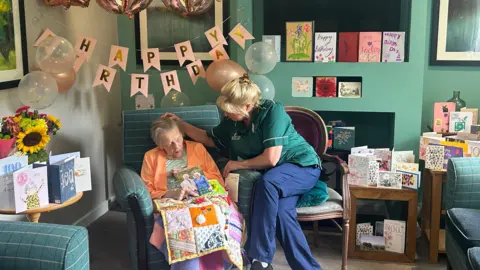 Bertha Preston sitting in a green arm chair in the care home, with a quilted blanket and a card from King Charles III resting on her lap. She is wearing a green top underneath an orange cardigan. Beside her is her carer, Jane Rogers, sitting with her legs crossed, wearing a green shirt and blue trousers. Jane is gently stroking the hair away from Bertha's face. They are surrounded by cards, happy birthday banners, balloons and flowers. 
