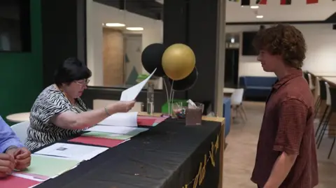 A boy in a brown shirt collects his A-Level results from a woman sat at a desk.
