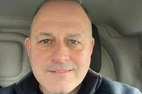 Mike Scourfield Man smiling in selfie style. He is balding and is sat in the car. He has blue/green eyes and is wearing a blue jumper. It is a head and shoulders shot.