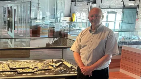 Amgueddfa Cymru Two men, one in his late 30s on the left and the other inhis late 60s on the right, stood in front of a display of treasure, including several historical gold coins from the reign of Ferdinand and Isabella of Spain, at the National Waterfront Museum in Swansea. 