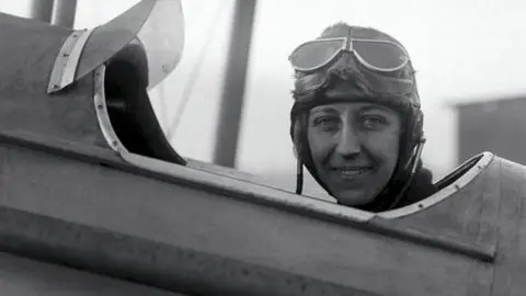 Black and white photograph of Amy Johnson dressed in flying gear in the cockpit of an aircraft. She is looking towards the camera and smiling, with her goggles pulled up on her forehead.