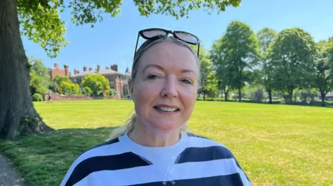 Louise pictured is looking into the camera and smiling. She's wearing a black and white striped sweatshirt, and has her sunglasses perched on the top of her head. She's stood in the shade underneath a large tree in a park, with bright green grass stretching away behind her. The sky is bright blue and clear, and there's a large, Victorian redbrick building on the horizon.