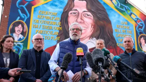 Reuters Gerry Adams stands in front of a huddle of microphones, with members of Sinn Féin standing behind him. He is speaking into the microphones and is wearing a blue gillet and a light blue shirt. He is standing in front of a mural of Bobby Sands.