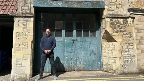 A man wearing a dark blue hoodie, black jeans and grey shoes stands in front of the facade of an old building, behind him a big blue wooden door which is shut. It's visible the building is in need of repair.