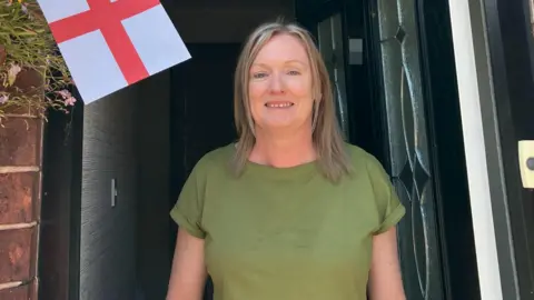 Maxine Simeone, who has shoulder length blonde hair and a green top, smiles at the camera from inside the doorway of a terraced house. A red and white England flag is to her right. 