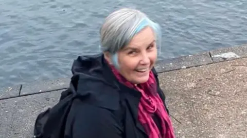 Pamela Chadwick stands beside a river or pond on a concrete path. She is smiling. She has short grey hair with streaks of blue and wears a pink scarf and navy coat. 