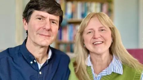 Portrait photo of Mark and Beverley Landreth-Smith, who have hosted seven different children over the last seven years. They are both smiling. Beverley is blonde with long hair and is wearing a green jumper over a blue and white striped shirt. Mark is wearing a navy blue shirt and has short, dark brown hair.