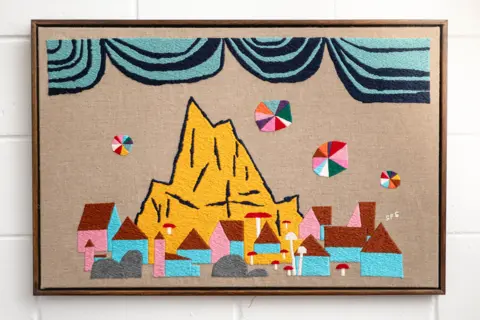 Stewart Francis Easton A stitched scene with a yellow mountain in the background of pink and blue buildings. A sky is depicted by blue striped waves. 