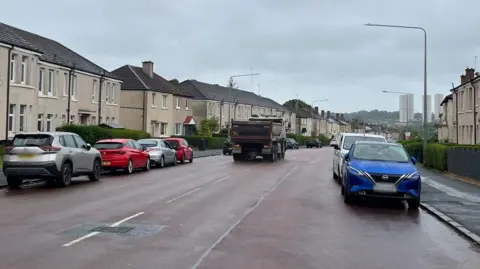 A street view of a wide main road with terraced homes on either side and vehicles parked along the pavement on both sides