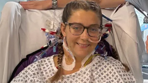 King's College Hospital NHS Foundation Trust Hannah Dyos in a hospital bed with various pieces of medical apparatus attached to her body. She is smiling and wearing glasses ad a hospital gown.