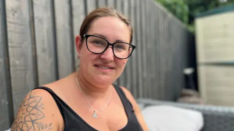 Head and shoulders image of Jade Gatt. Jade has brunette hair tied back, dark rimmed round glasses and a black sleeveless top. She is sitting on a garden chair with a fence behind her.