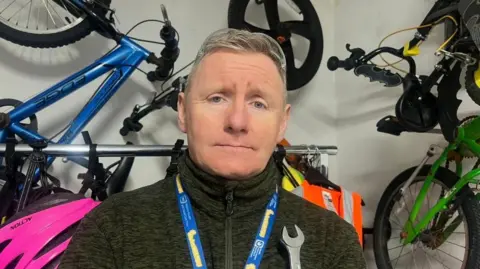 Danny Bryce is standing in a room with bikes mounted on the wall behind him. He has short grey hair swept to the side and is wearing a dark green fleece and a blue and yellow lanyard.