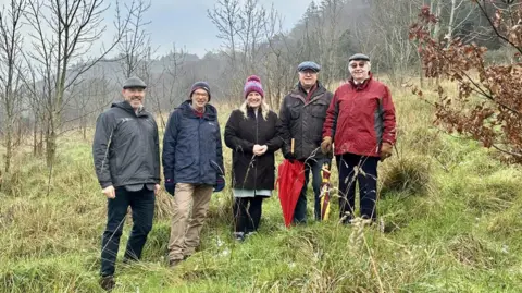 Five people standing beside each other smiling, on a plot of land which has been secured in the new deal. They are surrounded by trees which have no leaves.