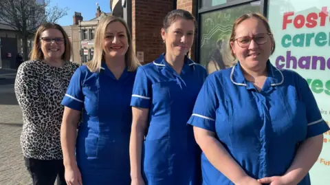 Four women standing in a line in front of a building. Three are wearing blue uniforms, the other has a white top with a brown pattern on it. They are all looking towards the camera and smiling.