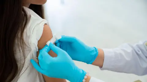 A girl with long dark hair seen from the back is getting a vaccine on her upper arm by a blue gloved hand.