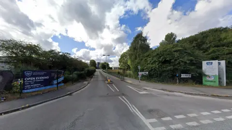 General view of the junction of a road with hedges and trees on both sides. On the left there is a dark blue banner with white writing on it. On the right, near a road sign, there is a white and green banner that also has writing on it