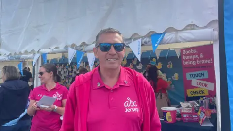 A white man in sunglasses and a pink t-shirt stands in front of an ABC Jersey breast cancer support stall.