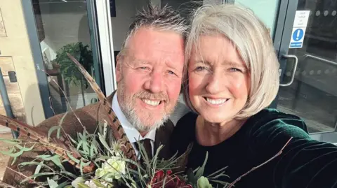 A bride and groom stand close together outside a modern building entrance, smiling at the camera. They hold a large, decorative bouquet filled with greenery, white and red flowers, and long feathers. 