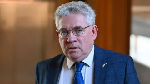 Kenneth Gibson, who has short grey hair and glasses, looks at the camera. He is wearing a blue suit and tie with a white shirt, with wood panelling in the background. 