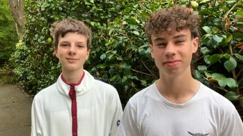 BBC/Seb Cheer Two teenage boys smile at the camera, standing in front of a hedge. A tree is also in the background.