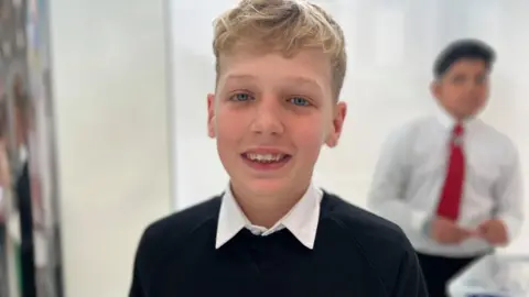 JJ looks at the camera. He has blond hair and is wearing a school uniform of a black jumper and white shirt - no tie