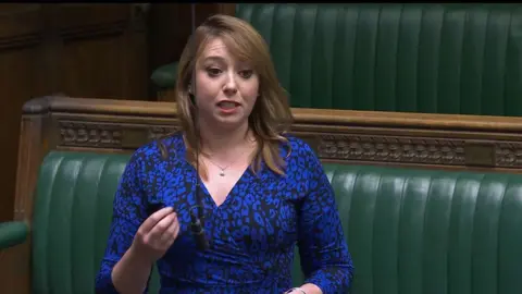 Sarah Bool with long brown hair wearing a blue dress with her right hand held out while making a point. She is standing in front of the green benches in the House of Commons.