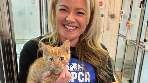 A woman with long blonde hair smiles at the camera while holding a ginger kitten