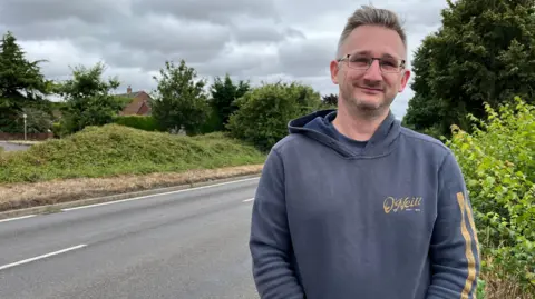 Clare Worden/BBC A man looks at the camera while wearing glasses and a navy sweatshirt. He is standing in front of a two lane road with bushes and vegetation either side.