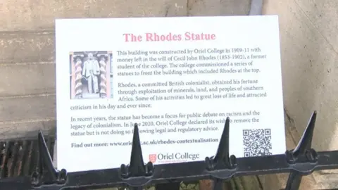 A white plaque explaining the history of Cecil Rhodes above some black fencing.