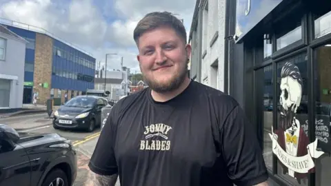 A man with a beard and brown hair smiling at camera. His t-shirt says Jonny Blades on the front. There are cars parked to his left and a barber shop to his right with a skeletal figure printed on the window.