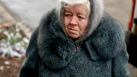 Goktay Koraltan/ BBC Lyuba stands outside wrapped up in a grey furry coat and hood with a sad expression on her face