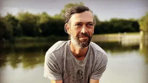 A photo of a man with grey hair and a grey beard, looking to the side of the camera. He is in front of a lake and many trees. He wears a grey T-shirt with a Bob Marley motif on it.