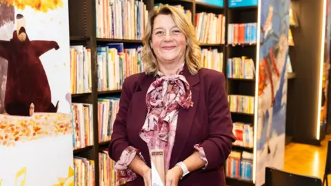 Adam Hollingworth Photography Julie Broadbent is stood in the middle of the image holding an award in the middle with both hands. She has shoulder length dark blonde hair and is wearing a pink blouse and burgundy suit jacket. She is stood in front of a book case. 
