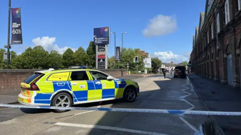 The cordon on Rodbourne Road, outside the Designer Outlet in Swindon. A marked police car is parked across the road, with investigative work happening in the distance.