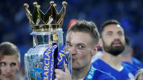 Action Images/Reuters A man in a blue football kit holds aloft a large silver trophy and kisses it while surrounded by team mates.