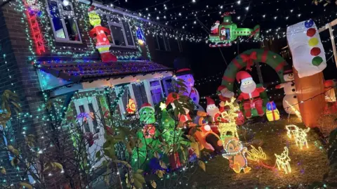 Green and white lights encompass a detached house with two floors with a blow up Grinch, helicopter, Santa, Christmas tree and snowman at the bottom. There are also light-up golden reindeers.